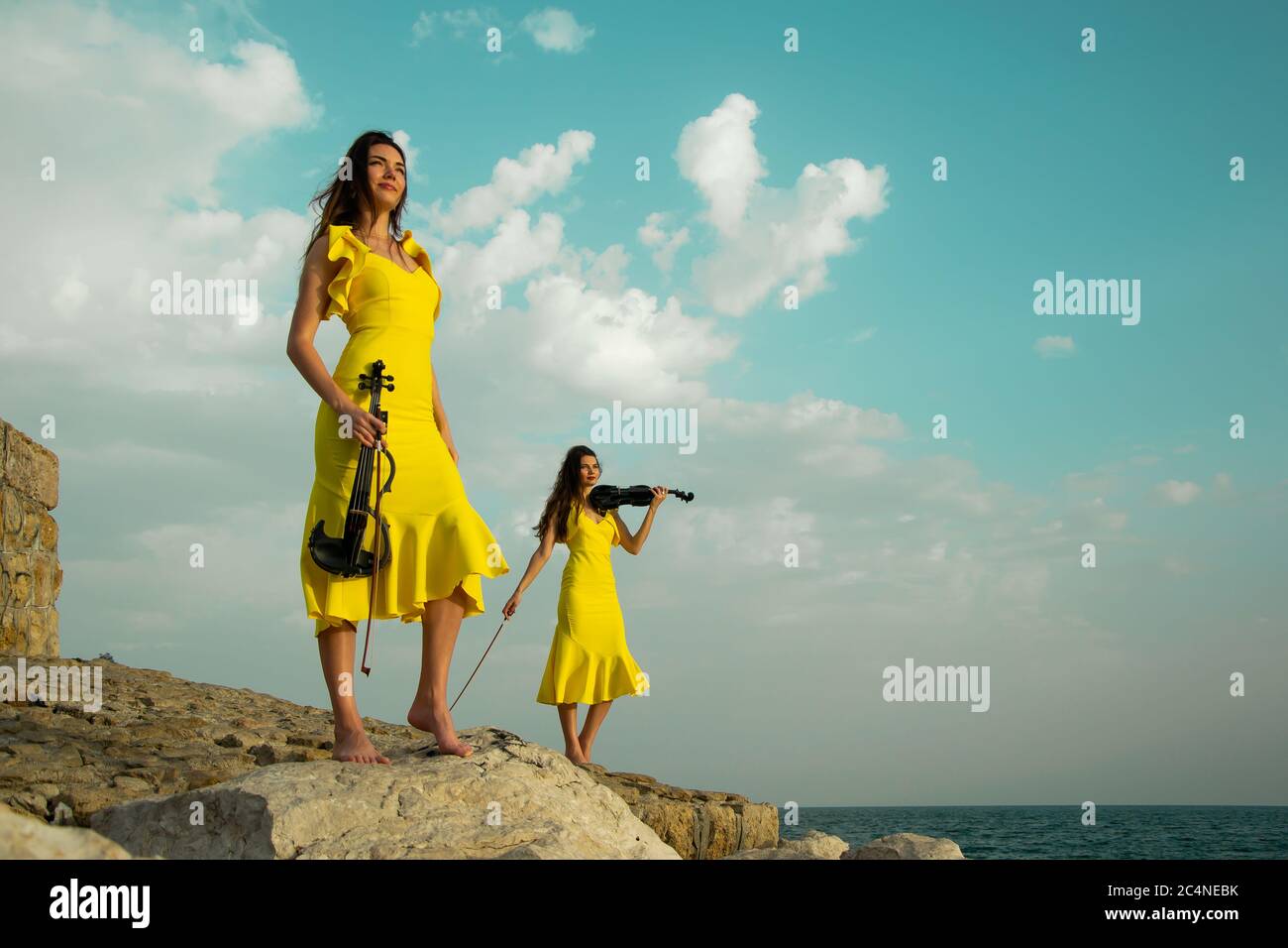 Deux belles sœurs jumelles violinistes dans des robes de concert jaunes jouent des violons électriques sur les rochers au bord de la mer Méditerranée à Antalya, Turquie. Mer Banque D'Images