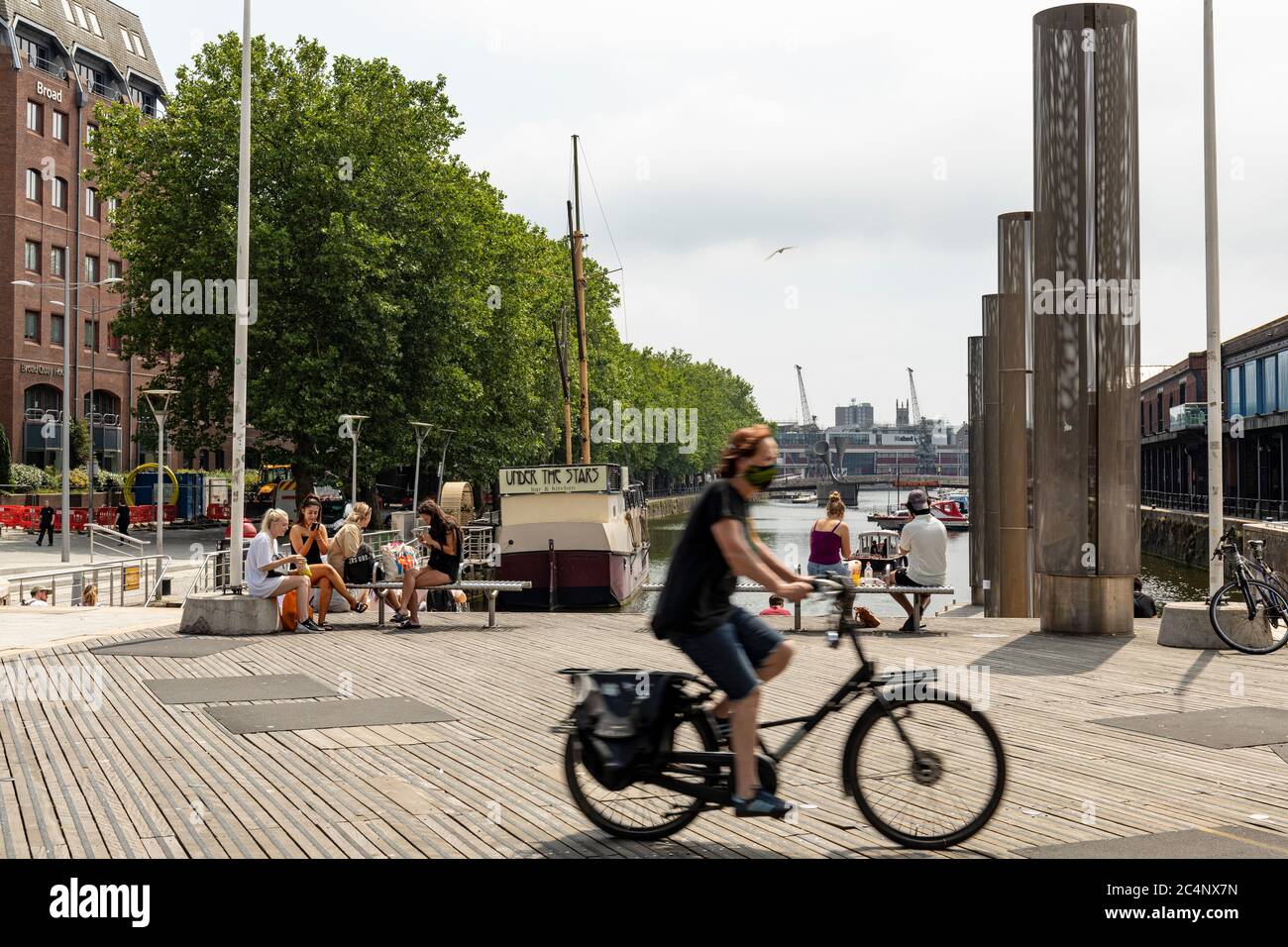 Après un relâchement des mesures de verrouillage un cycliste avec un masque de visage est en train de vélo au-delà des personnes appréciant le soleil à Bristol Harbourside. Bristol, Angleterre, Royaume-Uni Banque D'Images