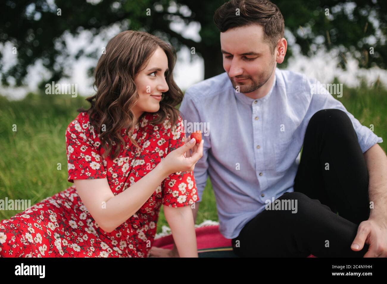 Jeune femme attirée avec un beau homme assis dehors sur une couverture rouge. Femme donner à l'homme fraise. L'homme goûte les baies Banque D'Images