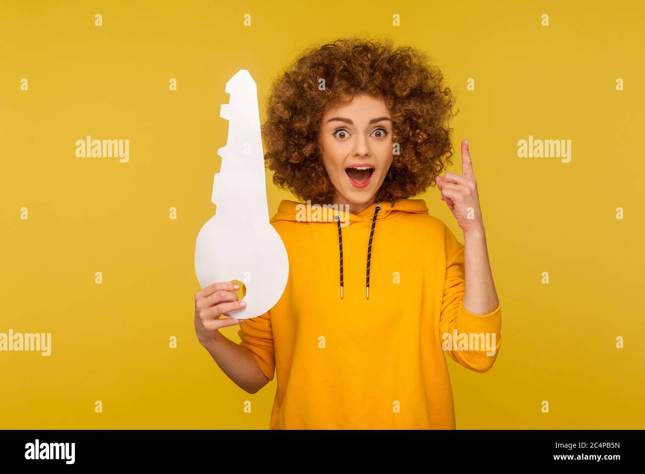 Portrait d'une femme inspirée et ébahi à cheveux bouclés dans un sweat à capuche de style urbain, montrant le doigt vers le haut avec une idée de génie et tenant une grosse clé en papier, semble surpris Banque D'Images