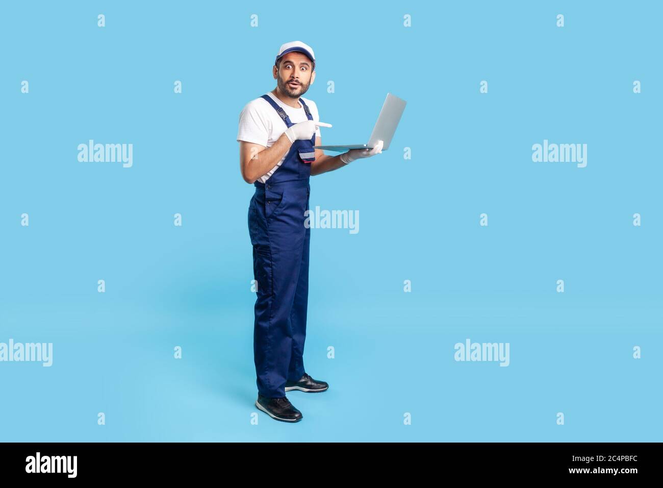 Pleine longueur stupéfait ouvrier en uniforme pointant vers l'ordinateur portable et regardant avec l'expression choquée, surpris par le message sur l'ordinateur, réparation ordre de service. Banque D'Images
