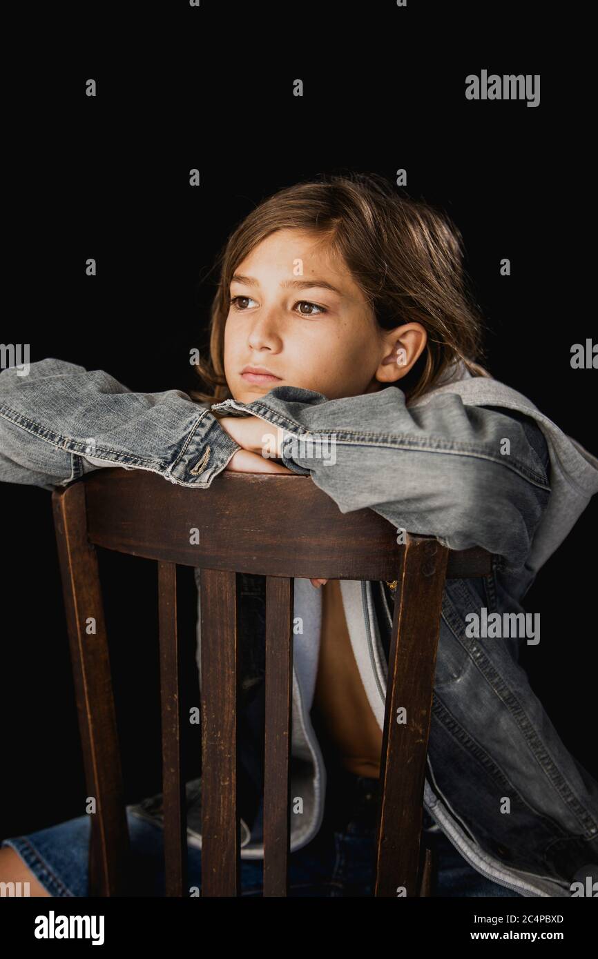 Garçon de 11 ans avec chandail à capuche et veste jean assis sur une chaise en bois sur fond noir Banque D'Images