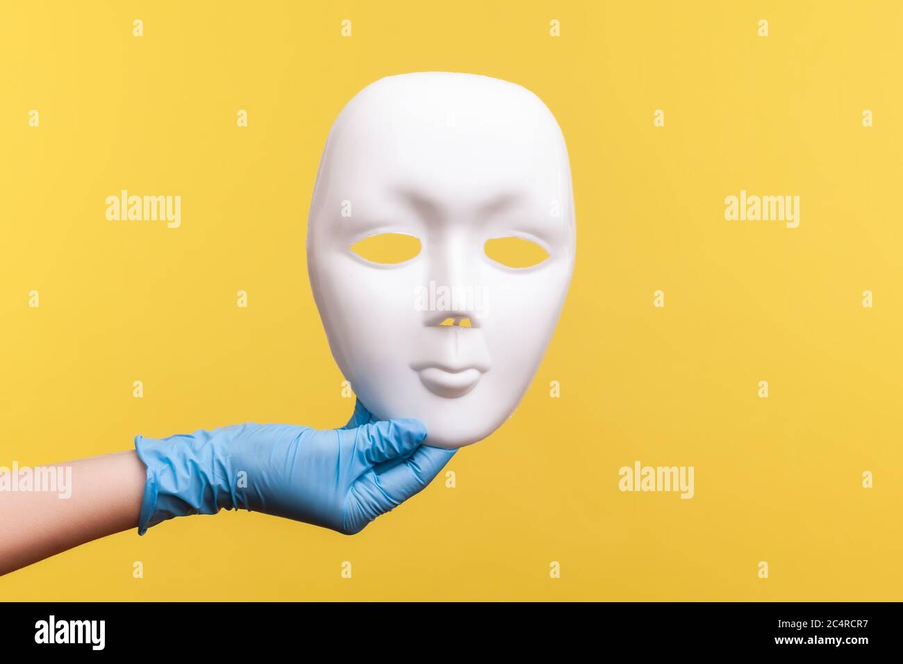 Vue latérale du profil gros plan de la main humaine avec des gants chirurgicaux bleus tenant un masque facial blanc. Prise de vue en studio, isolée sur fond jaune. Banque D'Images
