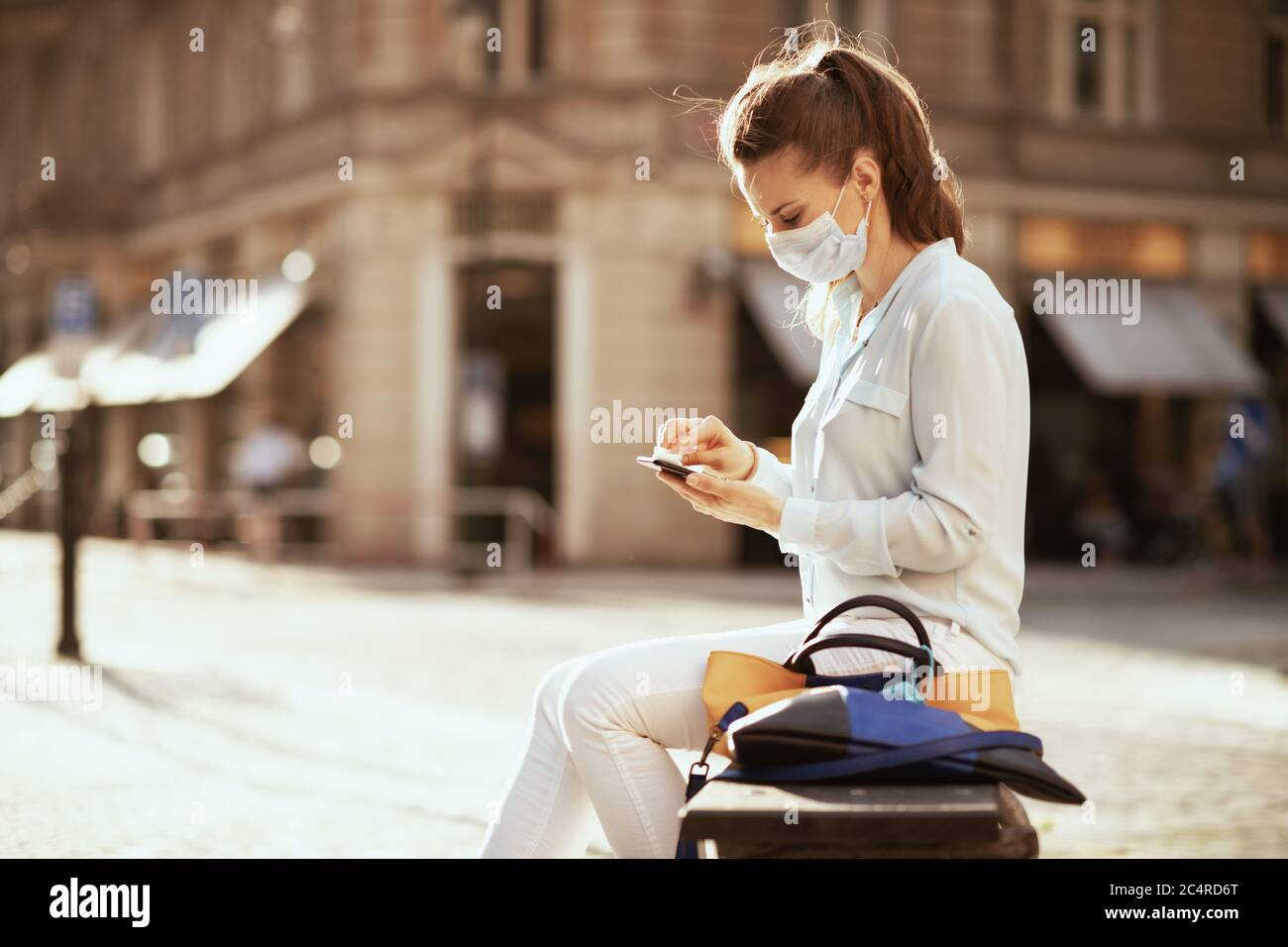 La vie durant la pandémie de Covid-19. Jeune femme en blouse bleue avec masque médical et sac à main smartphone essuyant avec assainisseur à l'extérieur de la ville. Banque D'Images