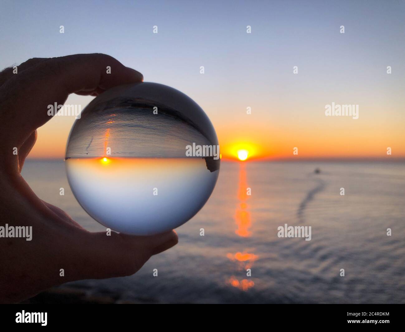 Main de femme tenant une boule de cristal, regardant à travers la mer et le ciel à l'aube. Concept créatif, voyage de rêve. Photographie de réfraction de boule de cristal Banque D'Images