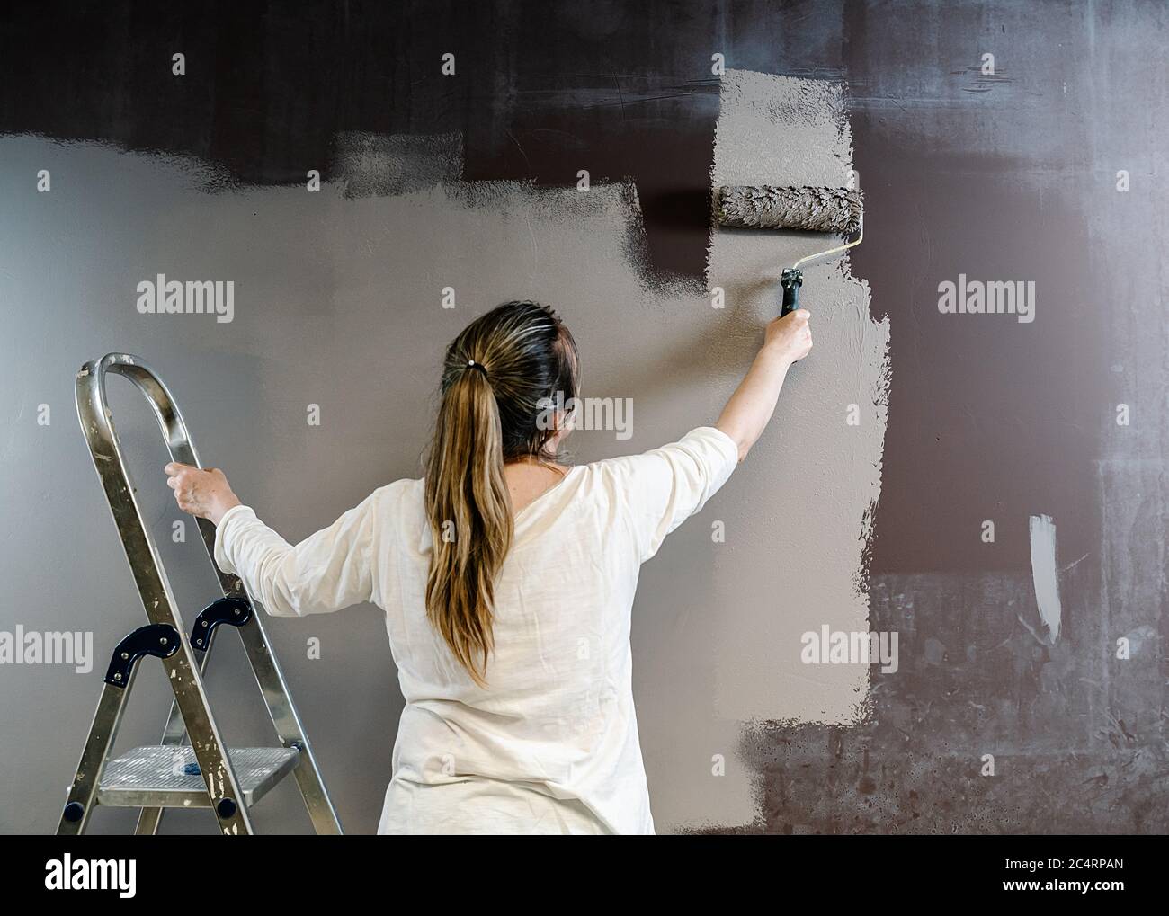 Une femme a grimpé une échelle et attrapé un rouleau de peinture plein de peinture grise sur un mur brun. Le peintre est en train de surépatiller et de faire passer le rouleau sur le mur Banque D'Images