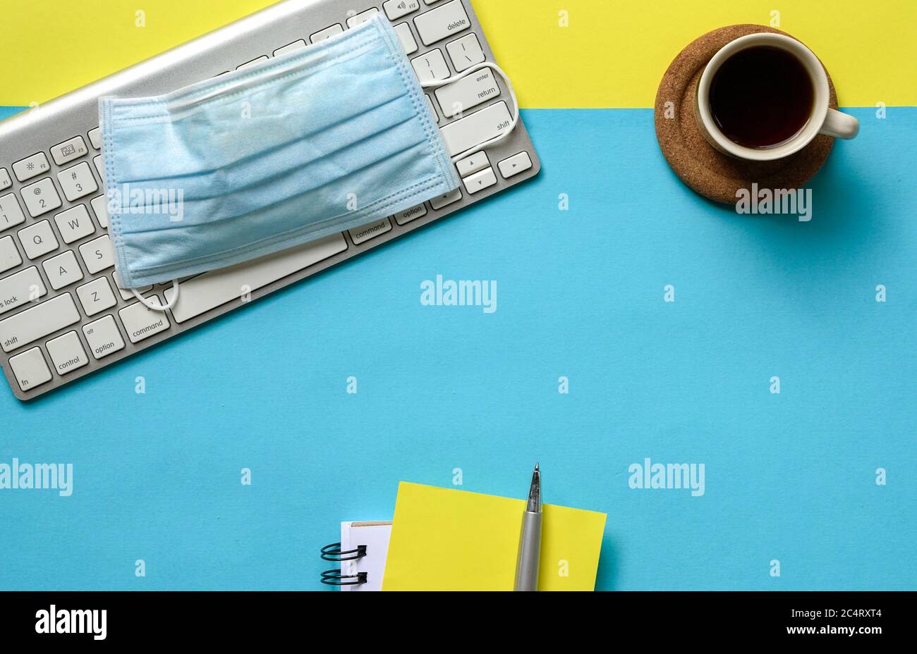 Plat de café, clavier sans fil, masque médical et carnet sur table jaune et bleue. Copier l'espace. Banque D'Images