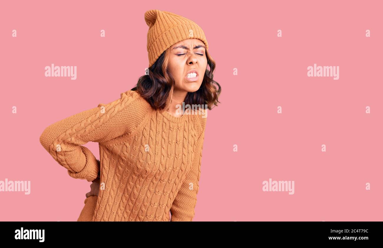 Jeune belle femme de course mixte portant un pull en laine et un chapeau  d'hiver souffrant de mal de dos, se touchant le dos avec la main, douleur  musculaire Photo Stock -