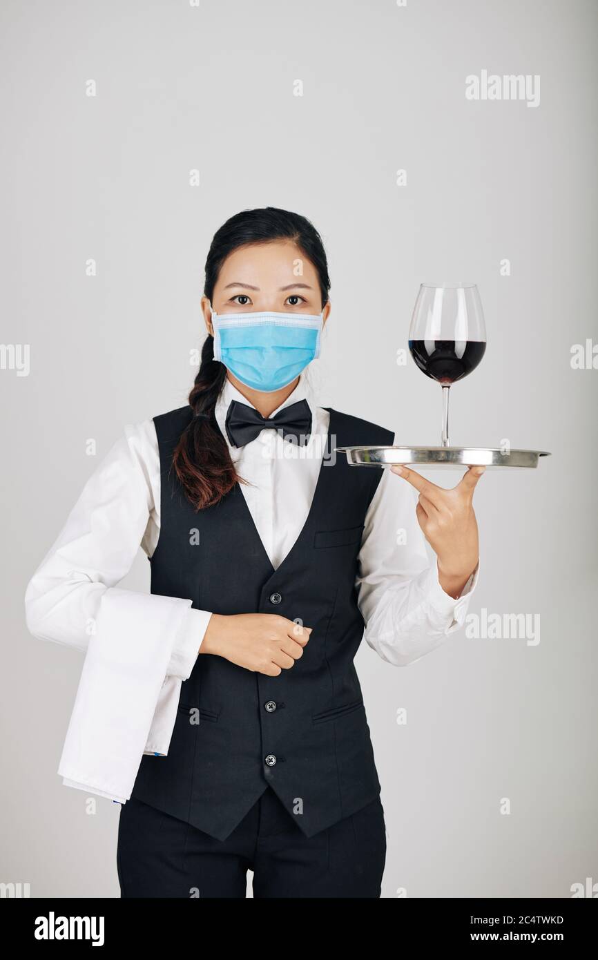 Jeune serveuse sérieuse portant une serviette blanche et un plateau avec un verre de vin, isolée sur le gris Banque D'Images