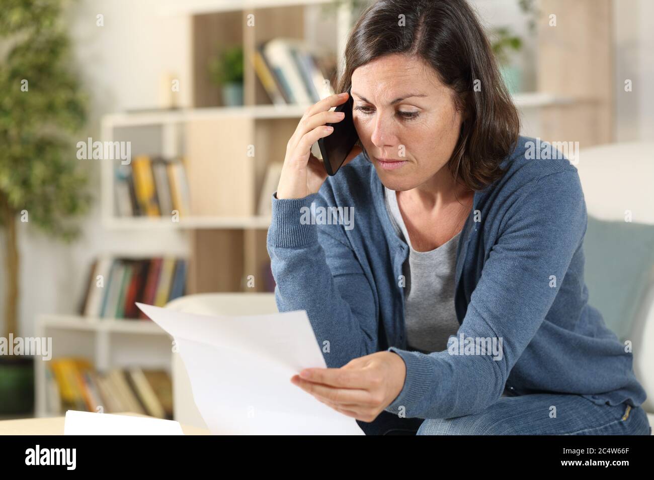 Femme adulte sérieuse appelant sur un smartphone vérifiant la lettre assise sur le canapé à la maison Banque D'Images