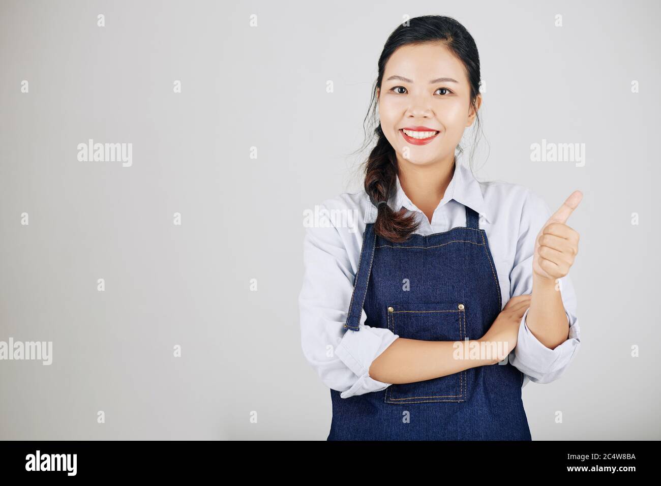 Souriant, joli jeune propriétaire d'entreprise en tablier en denim montrant le pouce et regardant la caméra Banque D'Images