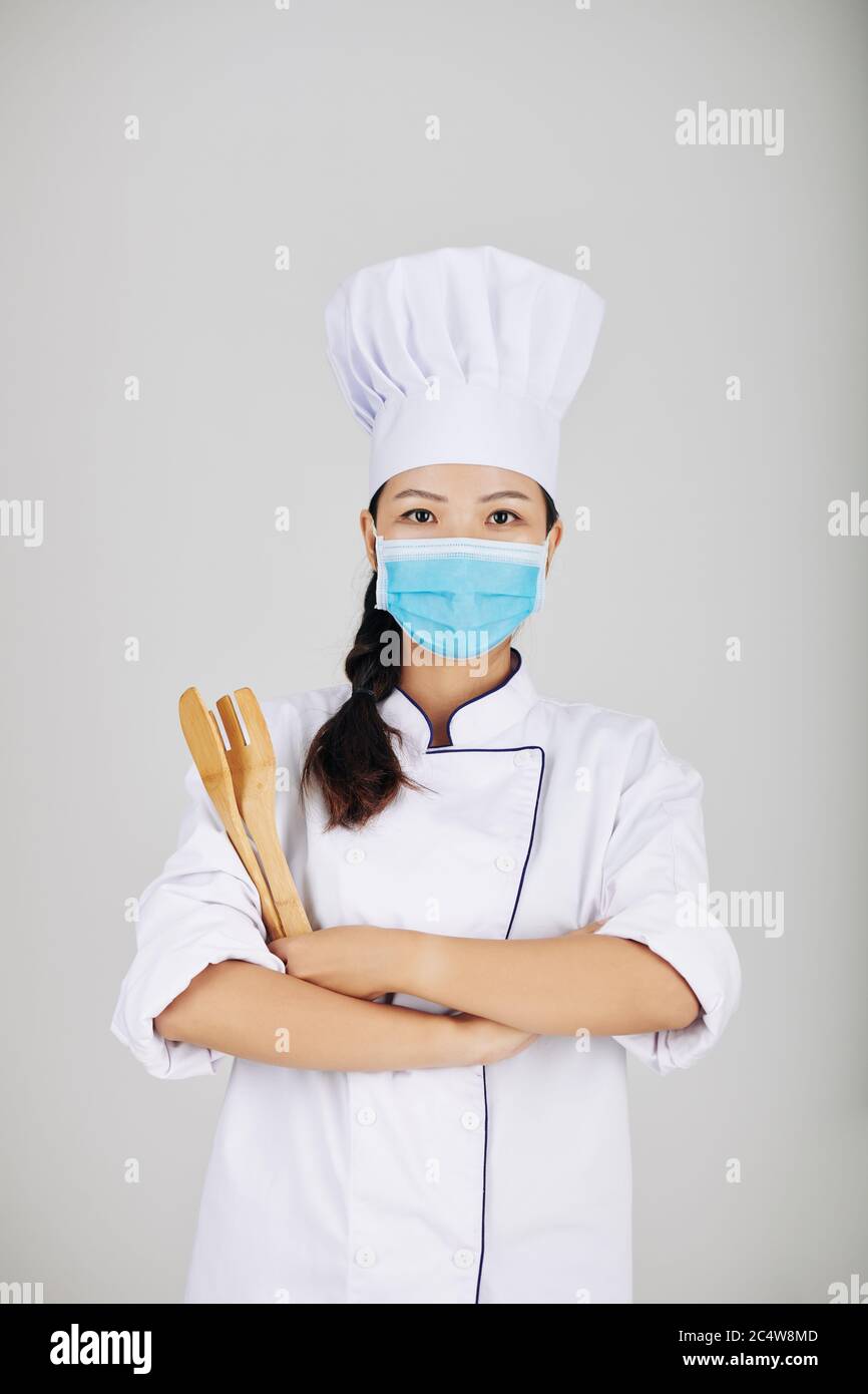 Portrait de jeune femme chef dans un masque médical tenant une fourchette et une spatule en bois lorsqu'elle pose contre le backgrounf gris Banque D'Images