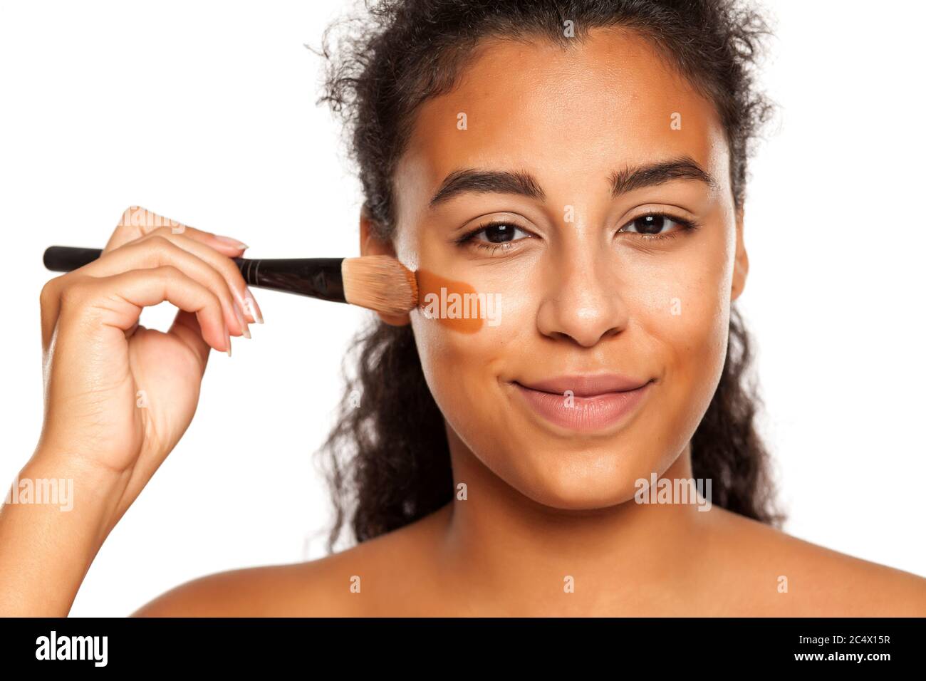 portrait d'une jeune femme à peau foncée appliquant un maquillage liquide avec une brosse sur son visage sur un fond blanc Banque D'Images