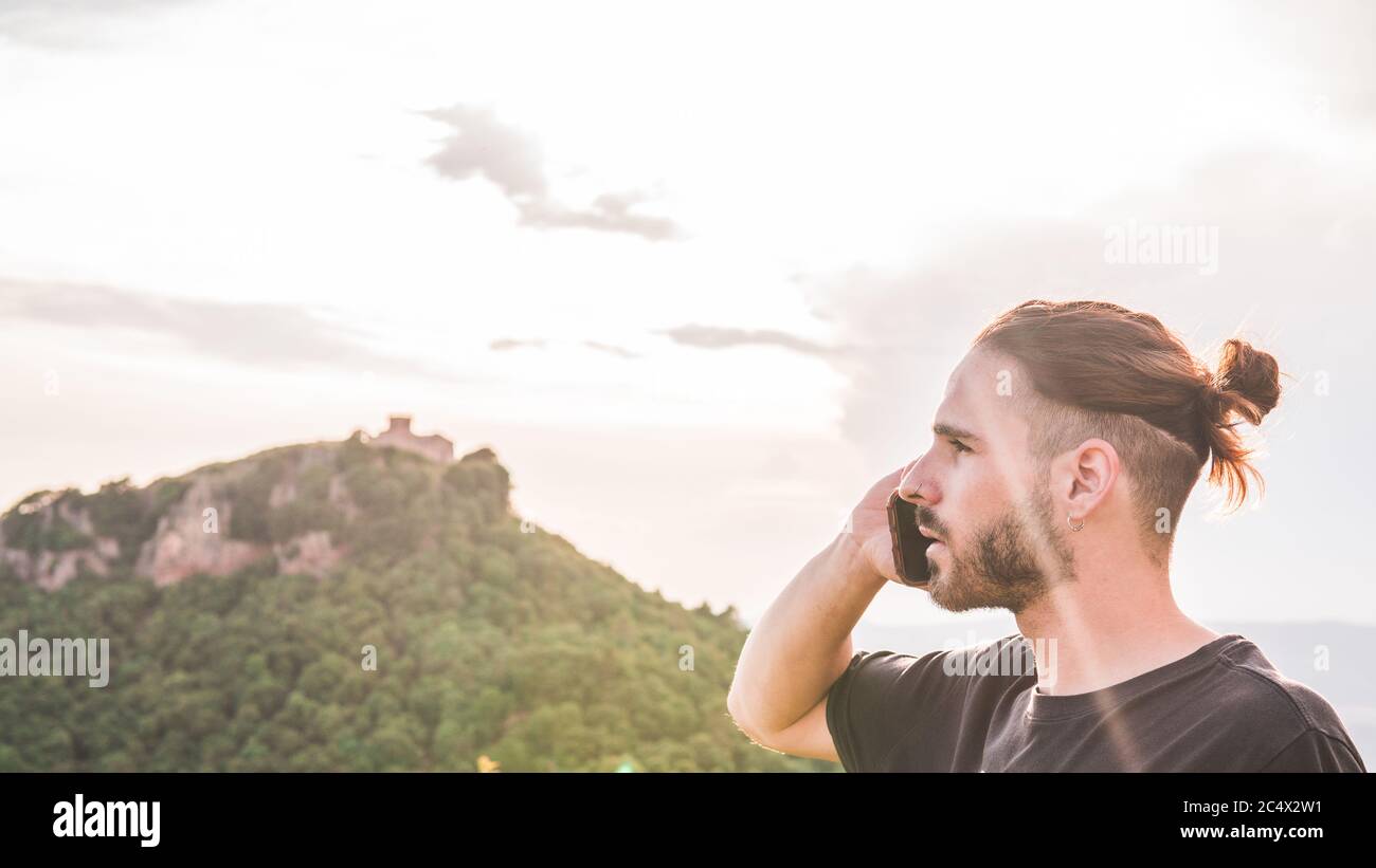 Un jeune homme beau dans le profil parle sur son téléphone cellulaire. Il y a une montagne avec un bâtiment au sommet. Banque D'Images