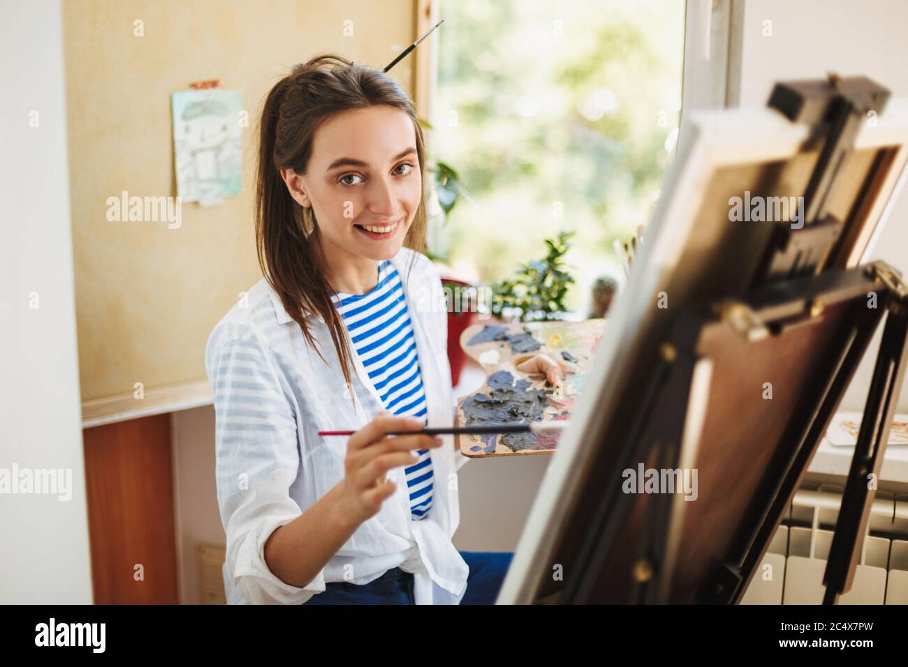 Jolie fille souriante en chemise blanche et T-shirt rayé tenant la brosse à peinture dans la main heureux de regarder dans l'appareil photo tout en dessinant sur le chevalet à la maison Banque D'Images