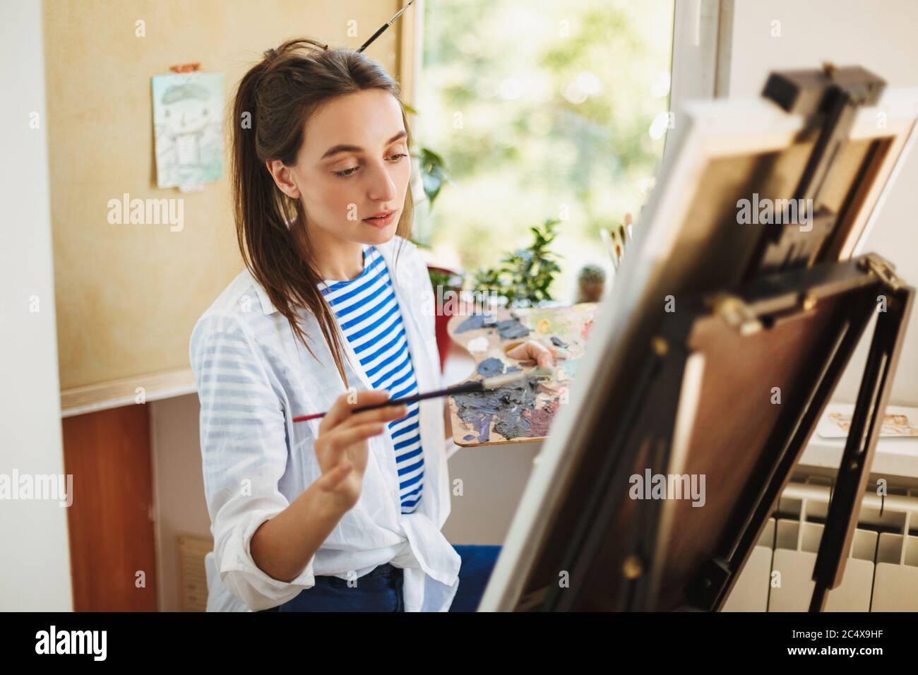 Une fille créative et réfléchie en chemise blanche et en T-shirt rayé tenant la brosse à peinture à la main tout en dessinant avec un chevalet à la maison Banque D'Images