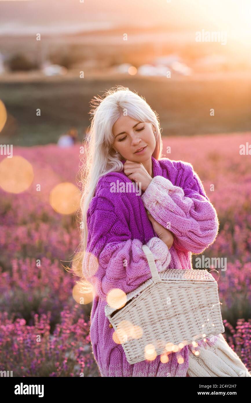 Blonde élégante femme 24-29 ans portant gilet tricoté élégant posant dans le champ de lavande cueillant des fleurs tenant panier de paille à l'extérieur. Été se Banque D'Images