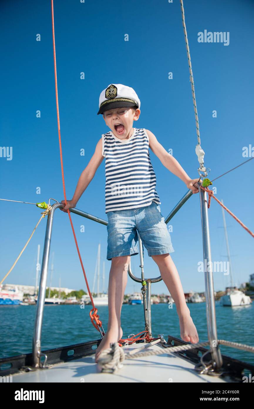 joyeux petit garçon mignon en capitaine chapeau blanc débardeur rayé et short en jean criant sur le yacht de luxe à la mer au port pendant le voyage de croisière d'été Banque D'Images