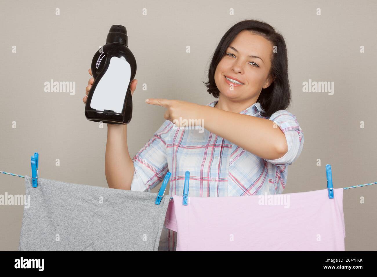 Belle femme avec bouteille de détergent en plastique noir suspendre chiffon humide propre pour sécher sur la ligne de vêtements à la buanderie sur un fond gris Banque D'Images