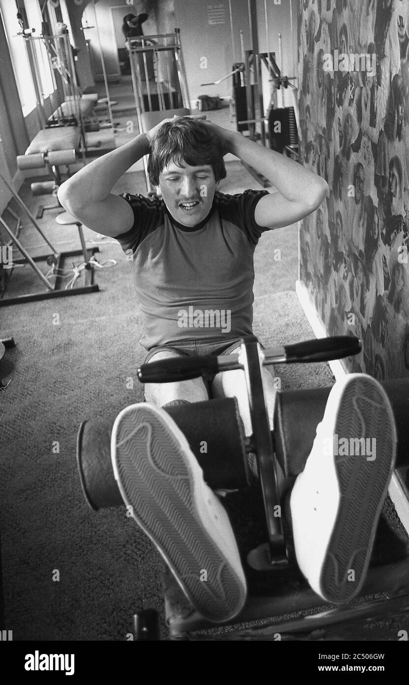 Années 1980, un homme dans une salle de gym allongé sur un banc avec ses pieds sous deux rouleurs faisant des sit-ups, Angleterre, Royaume-Uni. Les sit-ups sont un excellent exercice pour renforcer les muscles abdominaux, les fléchisseurs de hanche et le bas du dos. Fait correctement l'exercice peut construire le muscle et améliorer la posture. Banque D'Images