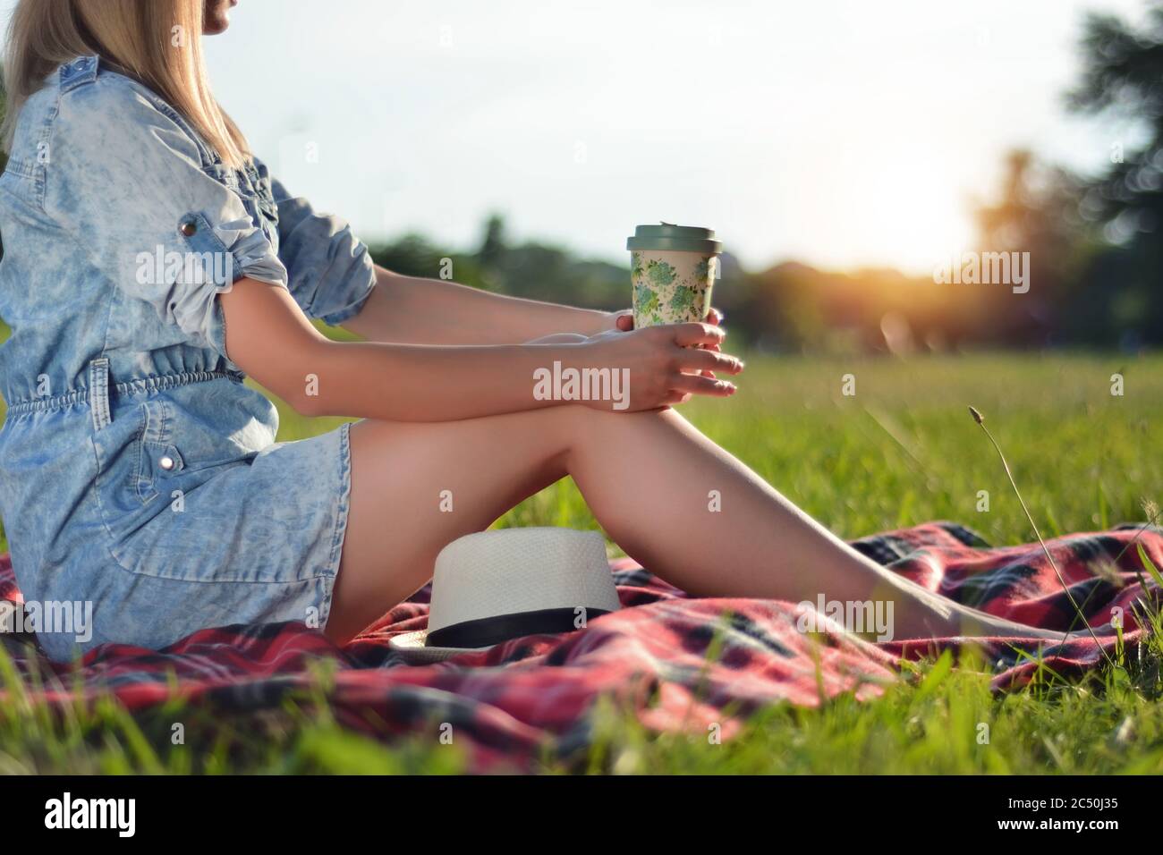 Femme avec une tasse de café sur les jambes assise sur une couverture rouge dans l'herbe verte de la prairie dans le parc et appréciant dans le coucher du soleil Banque D'Images