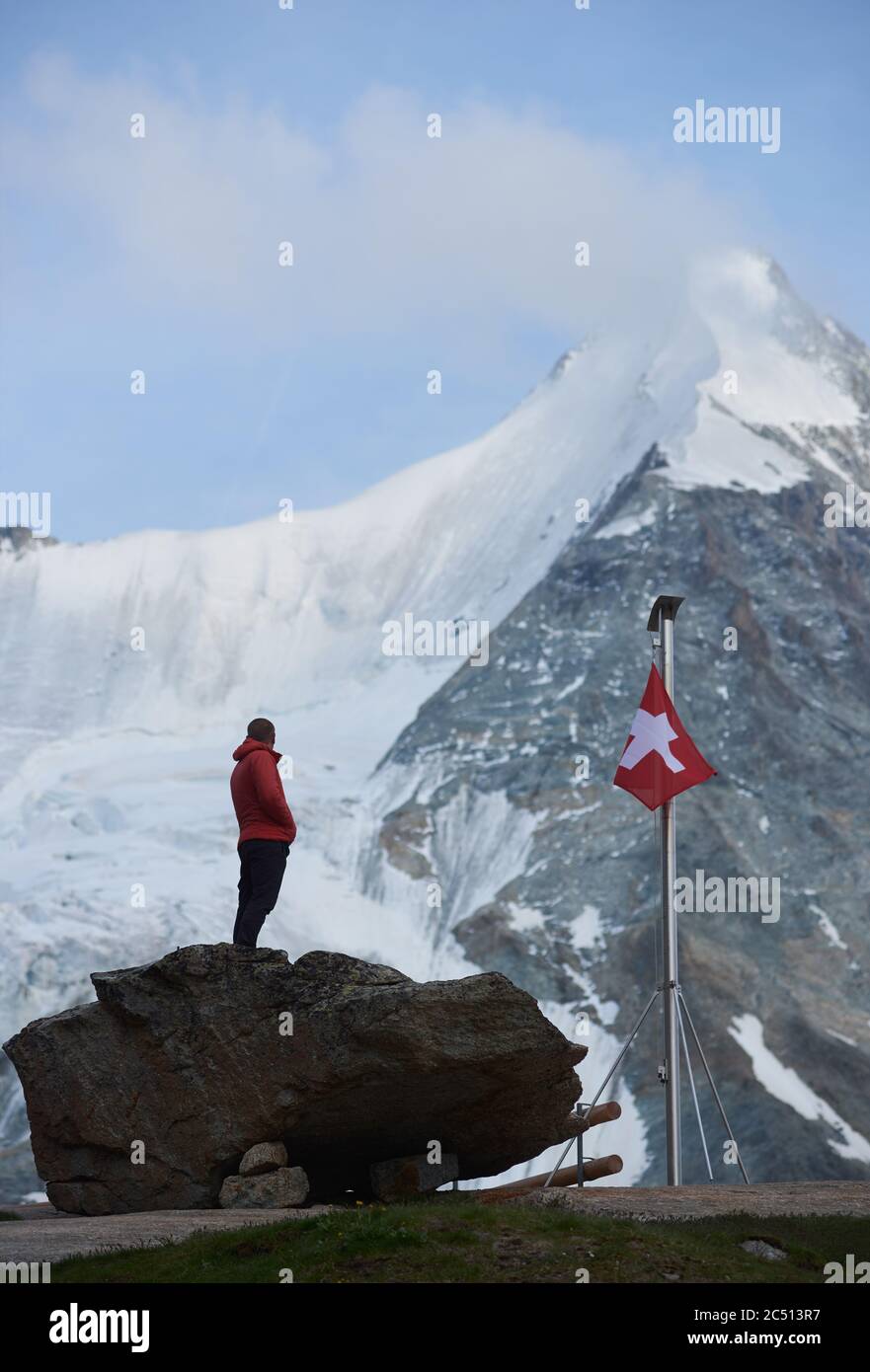 Mâle touriste regardant de beaux paysages de montagnes . Homme debout sur des pierres près du mât avec drapeau suisse. Ober Gabelhorn est en arrière-plan. Nature sauvage avec vues incroyables. Tourisme dans les Alpes. Banque D'Images