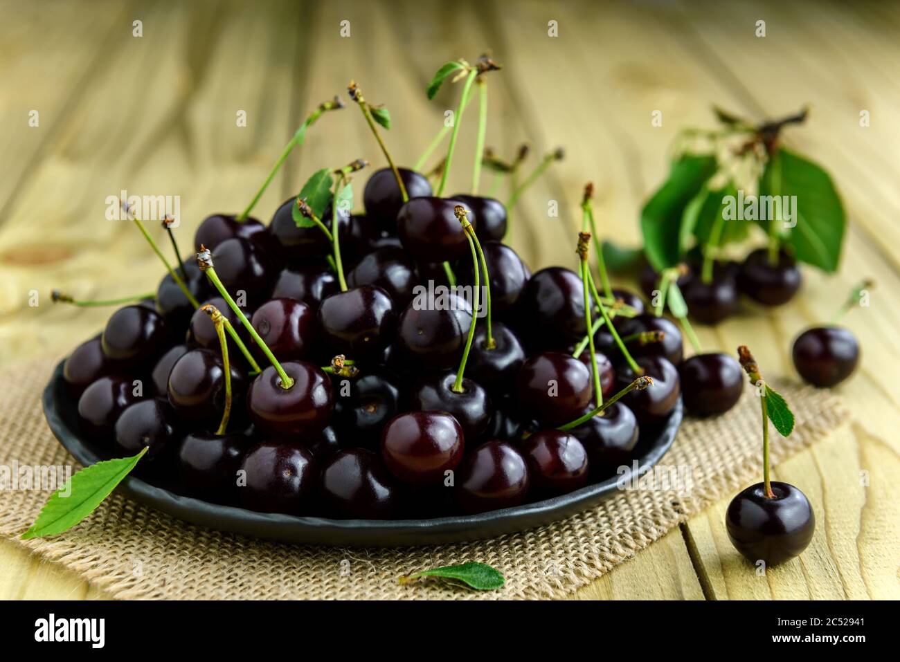 Cerises fraîches sur une assiette noire et feuilles vertes sur la table en bois. Cerises fraîches mûres. Mise au point sélective. Banque D'Images