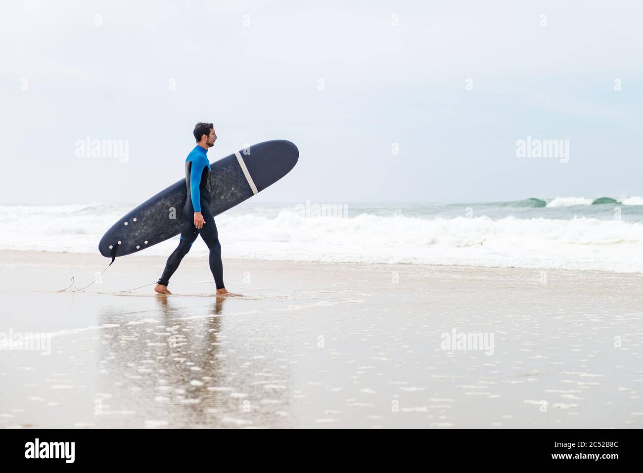 Un jeune surfeur mâle portant une combinaison, tenant une planche de surf sous son bras, marchant sur la plage après une séance de surf matinale. Banque D'Images