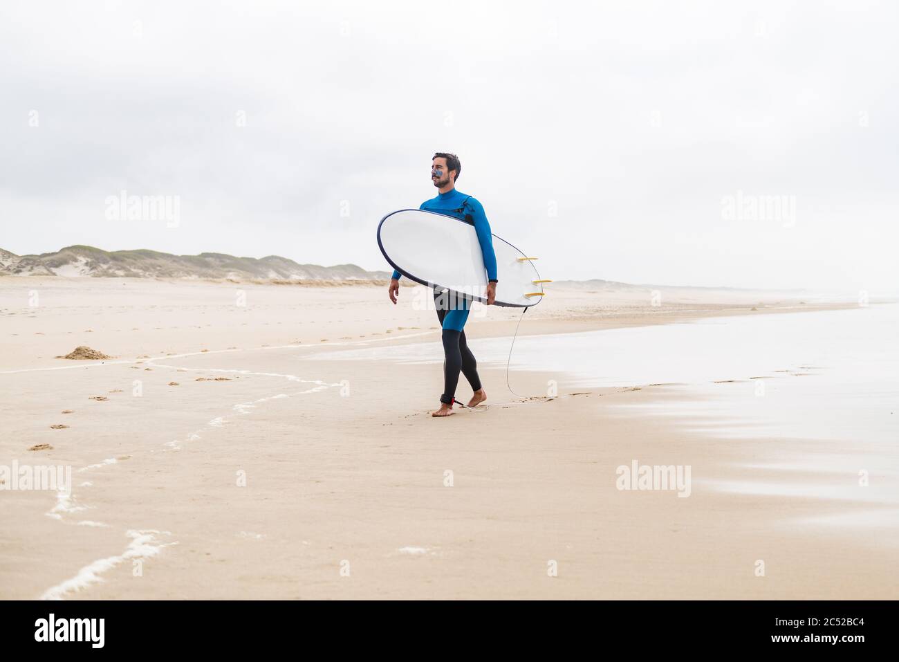 Un jeune surfeur mâle portant une combinaison, tenant une planche de surf sous son bras, marchant sur la plage après une séance de surf matinale. Banque D'Images