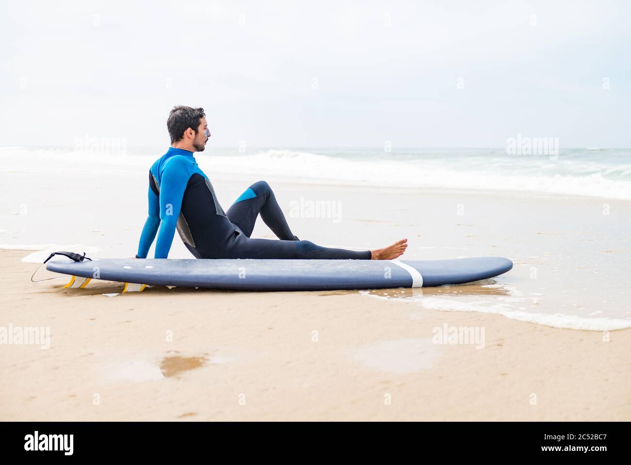 Jeune surfeur masculin portant une combinaison, assis à côté de son surf sur la plage après une séance de surf le matin. Banque D'Images