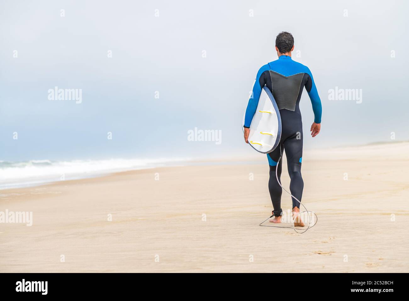 Un jeune surfeur mâle portant une combinaison, tenant une planche de surf sous son bras, marchant sur la plage après une séance de surf matinale. Banque D'Images