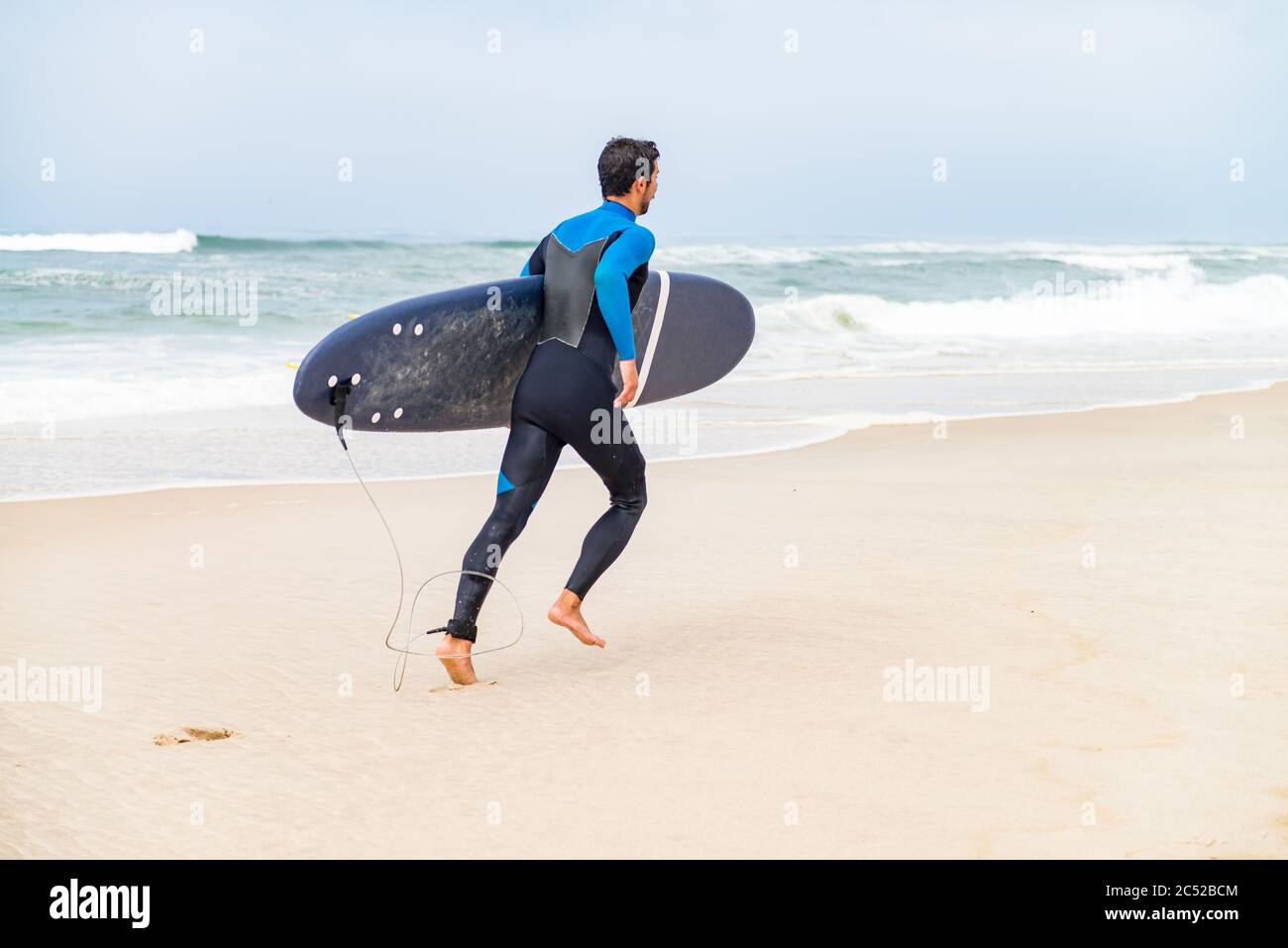 Un jeune surfeur masculin portant une combinaison, tenant une planche de surf sous son bras, courant sur la plage après une séance de surf matinale. Banque D'Images