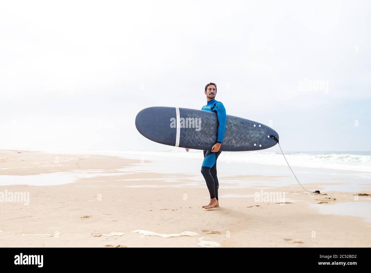 Jeune surfeur mâle portant une combinaison, tenant la planche de surf sous son bras, debout sur la plage après une séance de surf matinale. Banque D'Images