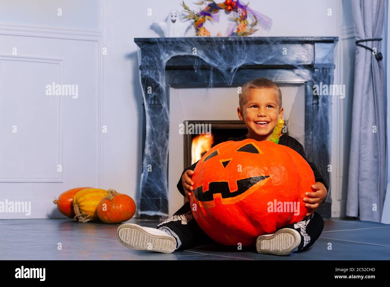 Petit garçon caucasien souriant avec panneau de trick ou de traite et grand citrouille d'Halloween orange assis près de la cheminée Banque D'Images