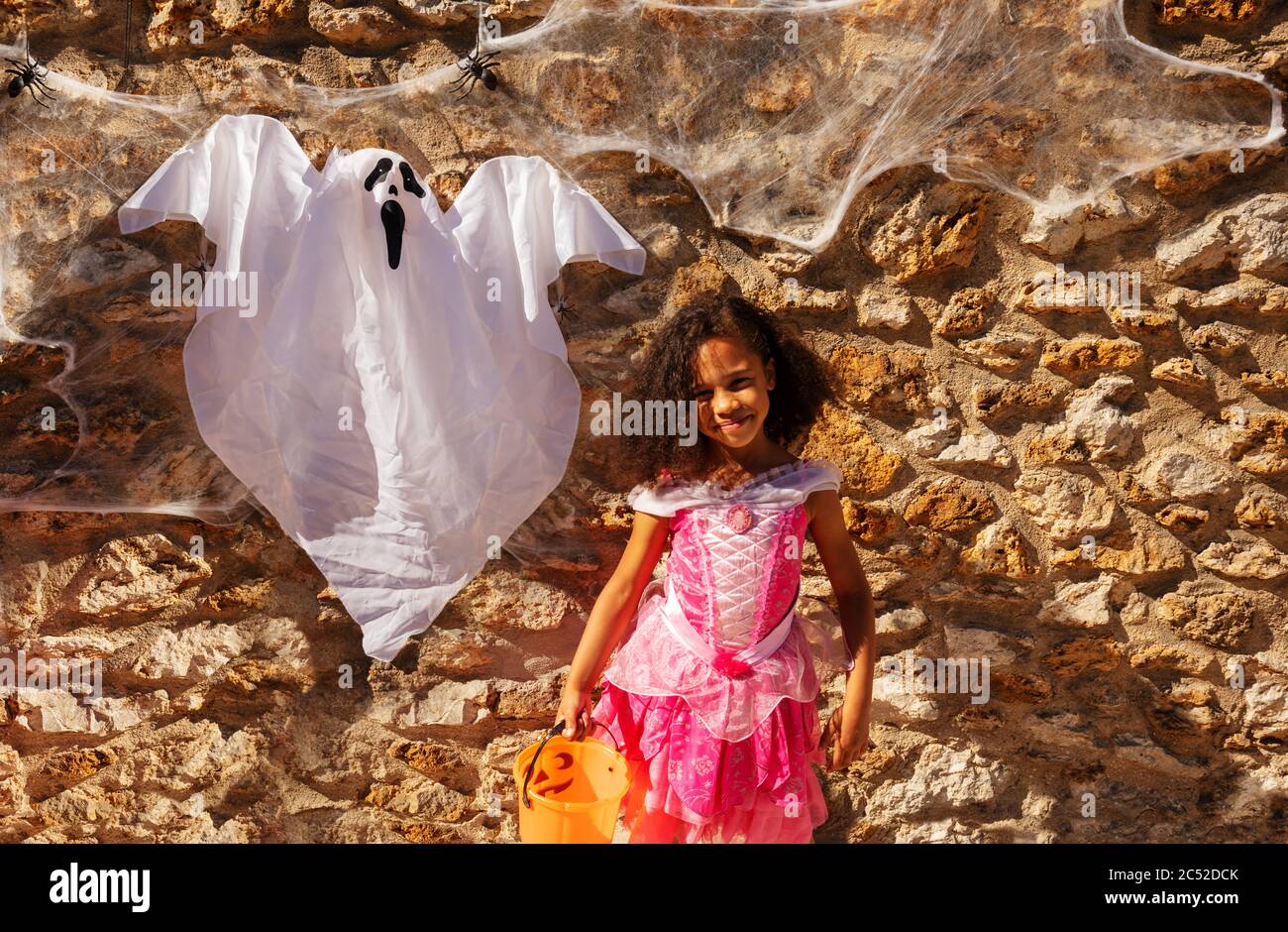 Portrait de la jolie fille curly dans le stand de costume d'Halloween tenant le seau pour les bonbons près du mur avec le fantôme Banque D'Images