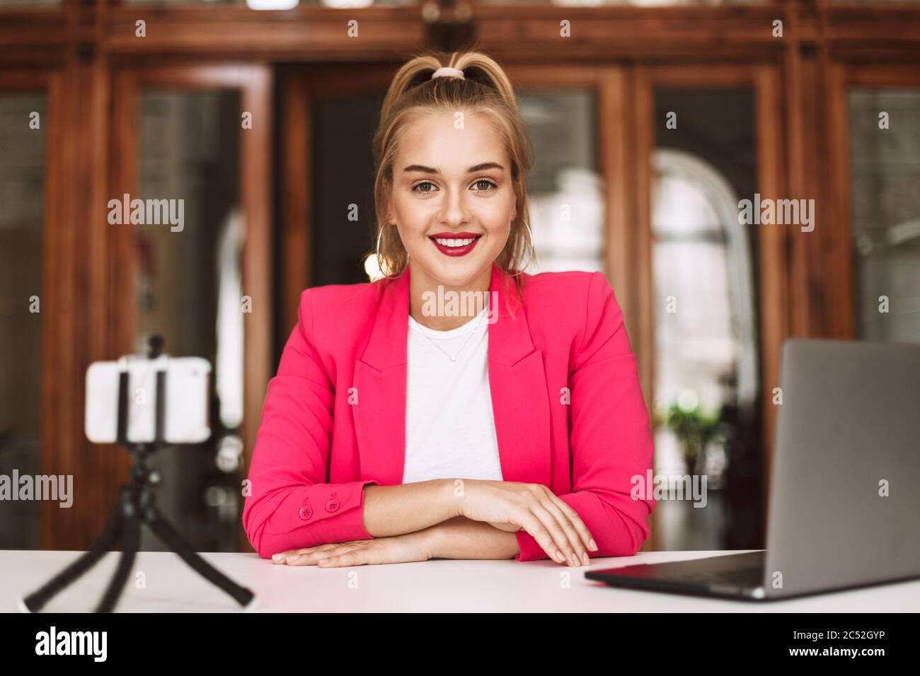 Belle fille souriante dans la veste rose heureux regardant dans l'appareil photo tout en enregistrant de nouvelles vidéos avec le téléphone portable et l'ordinateur portable Banque D'Images