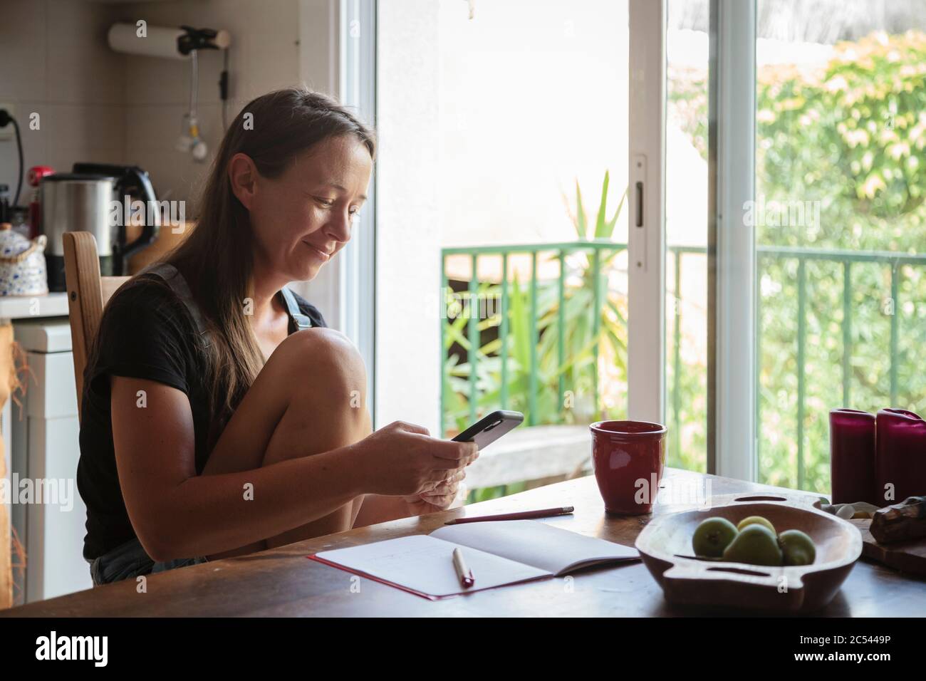 Jeune femme assise sur une table de cuisine en bois avec une atmosphère détendue en vérifiant son smartphone et en écrivant des notes; concept de style de vie naturel, simplicité Banque D'Images