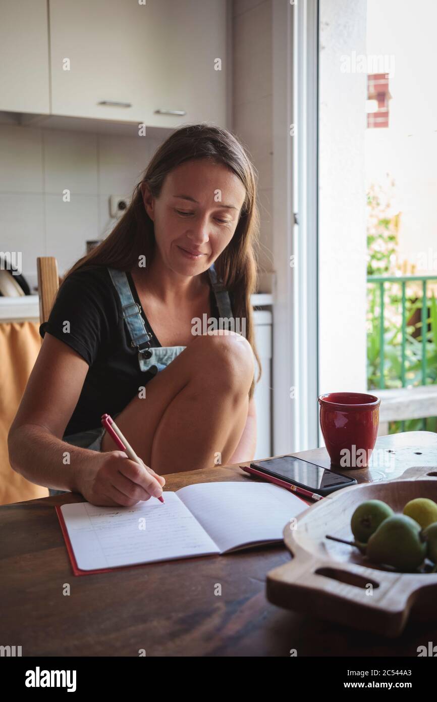 Jeune femme assise sur une table de cuisine en bois avec une atmosphère détendue en vérifiant son smartphone et en écrivant des notes; concept de style de vie naturel, simplicité Banque D'Images