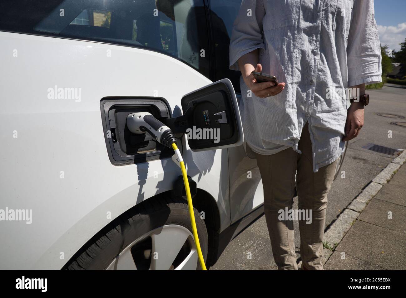 Une femme charge une voiture électrique blanche avec un téléphone portable dans elle main Banque D'Images