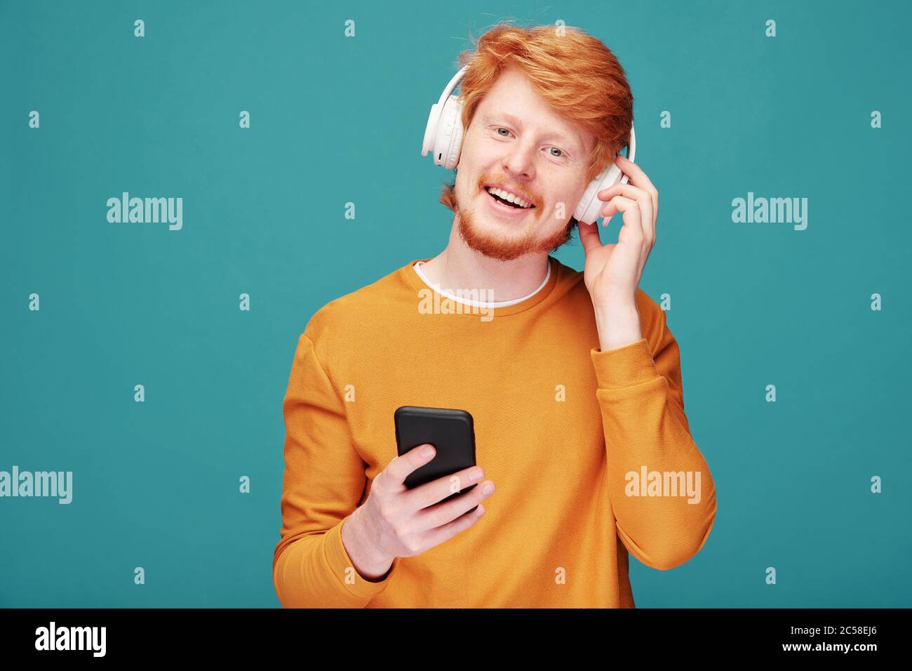 Portrait d'un jeune homme à tête rouge, beau et positif, avec barbe, réglage du casque sans fil tout en choisissant la piste musicale sur un smartphone Banque D'Images
