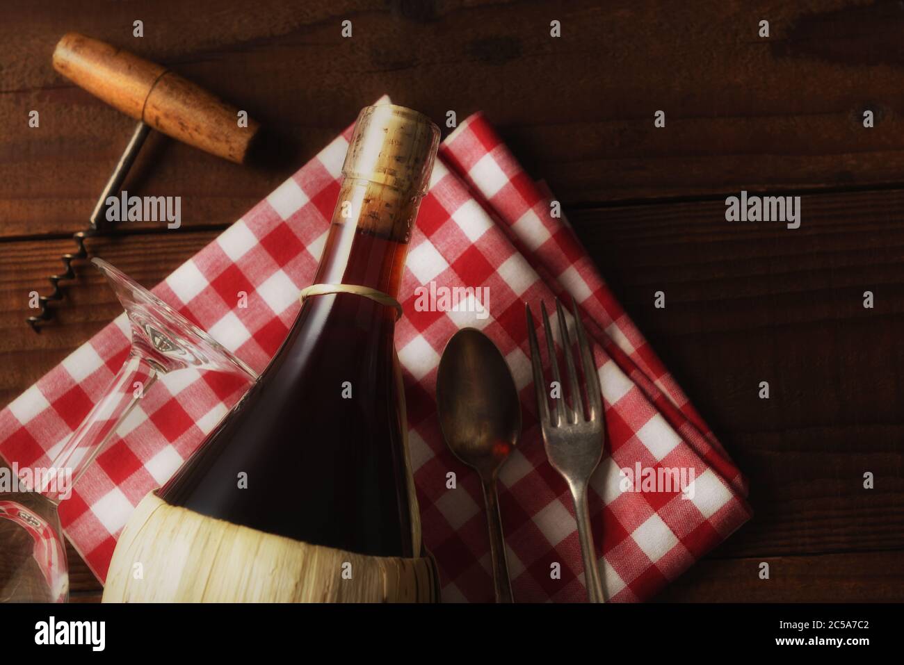 Flat Lay Wine STILL Life : un panier de vin Chianti sur une serviette à carreaux rouge et blanc avec tire-bouchon, fourchette et cuillère avec lumière d'ambiance chaude. Banque D'Images