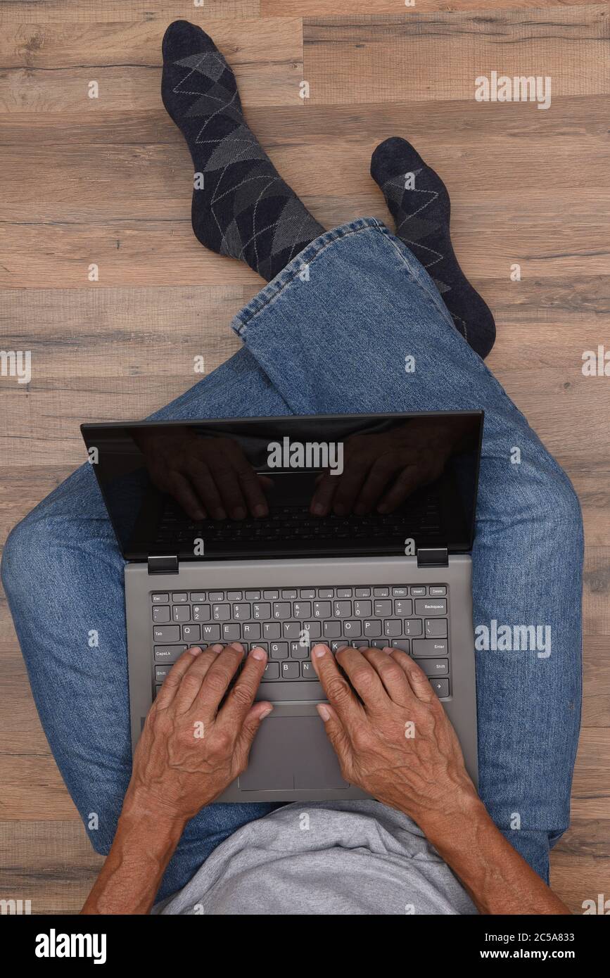 Concept de la maison du formulaire de travail. Angle élevé d'un homme assis sur le sol avec son ordinateur portable. La personne méconnaissable est vêtue de bleu denim Banque D'Images