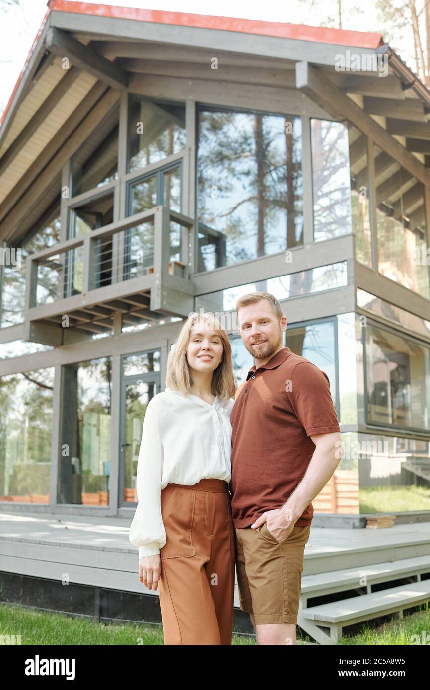 Portrait d'un jeune couple souriant en tenues décontractées debout contre une maison en verre et s'embrassant Banque D'Images