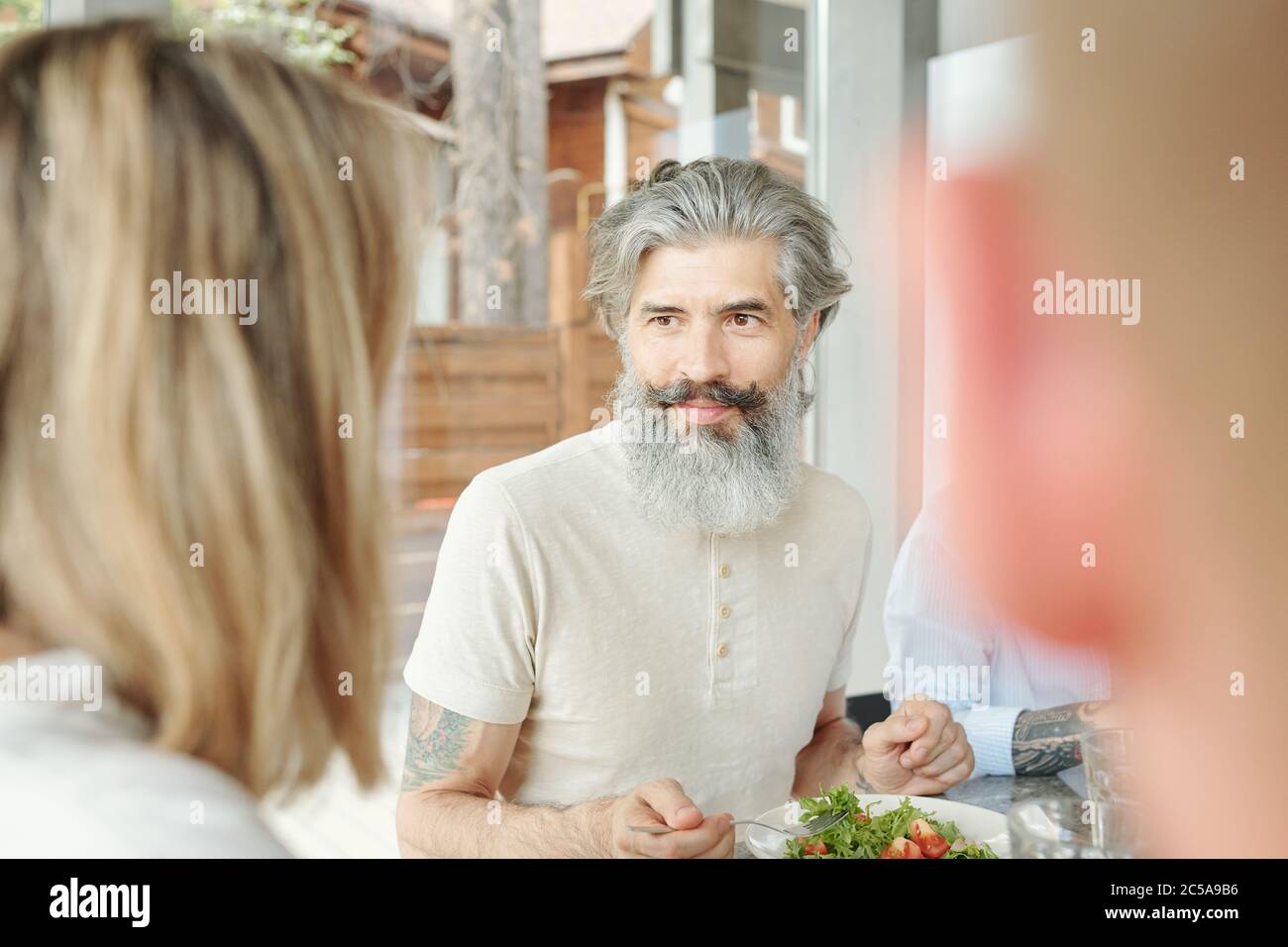 Beau homme âgé avec moustache et barbe assis à table et en train de manger de la salade tout en parlant aux clients au dîner Banque D'Images