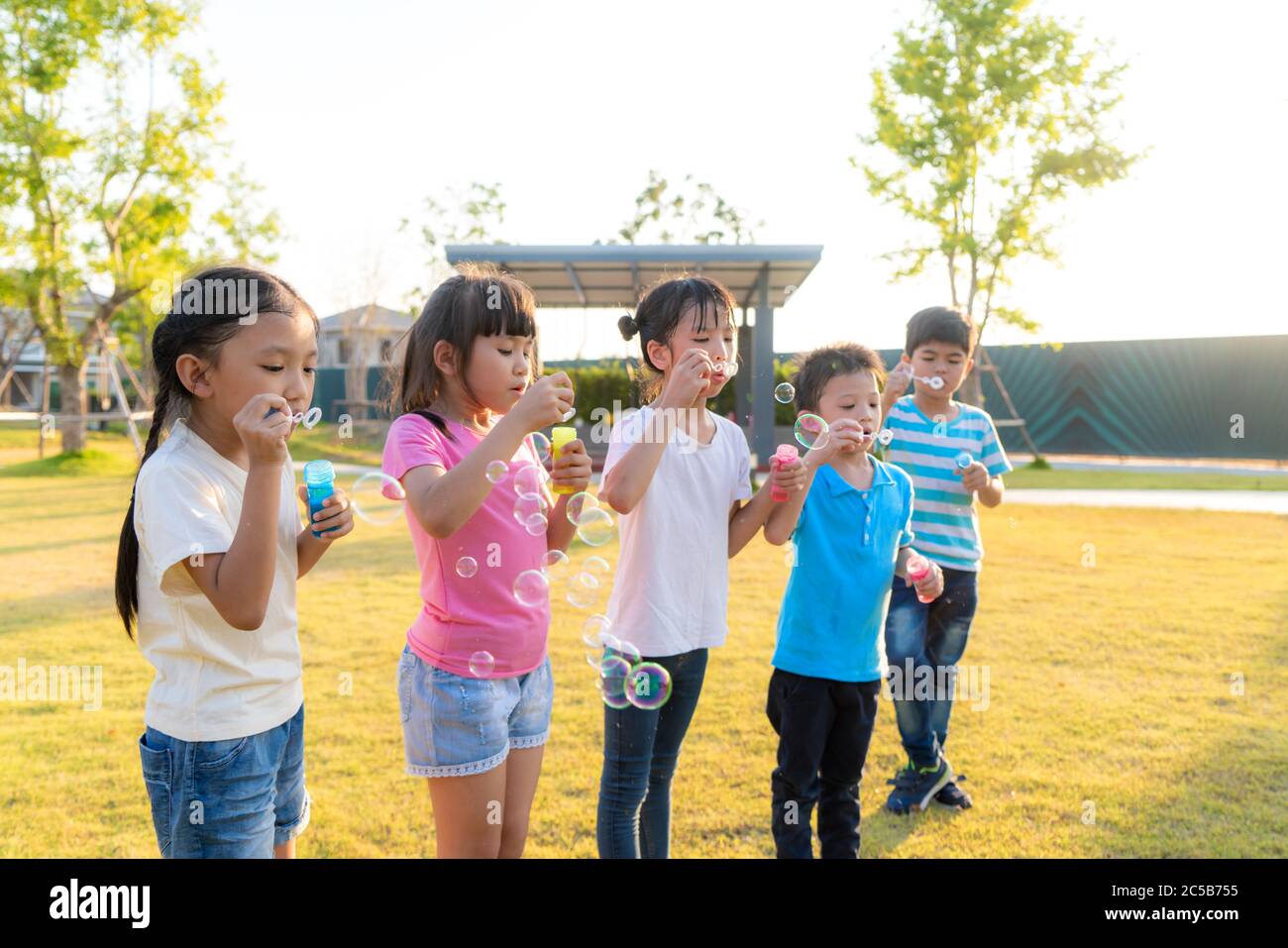 Un grand groupe d'heureux amis asiatiques souriants de maternelle gamins jouant des bulles de souffle ensemble dans le parc sur l'herbe verte le jour ensoleillé d'été. Banque D'Images