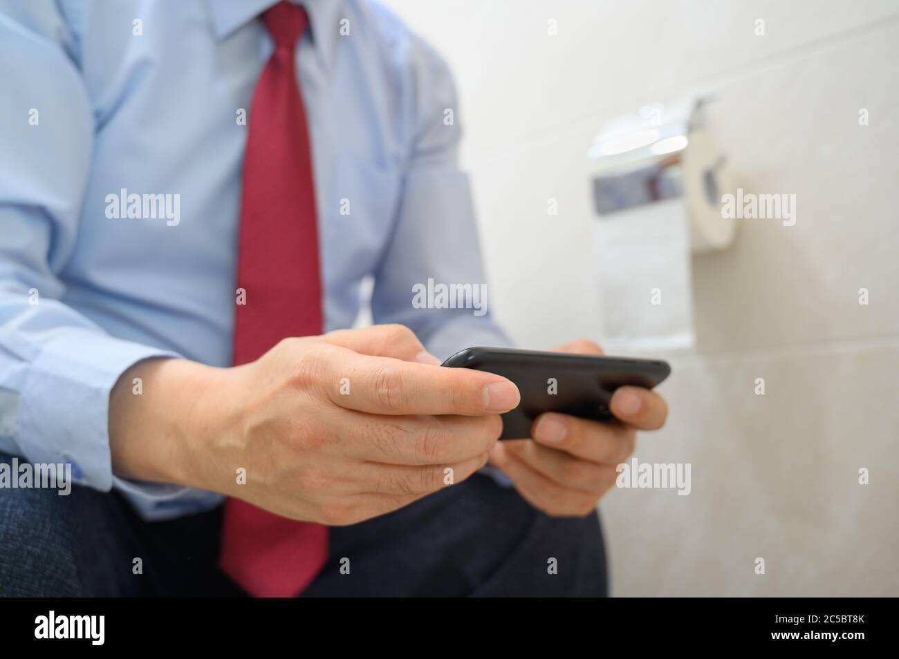 Homme d'affaires assis sur les toilettes dans la salle de bains et à l'aide d'un smartphone. Banque D'Images