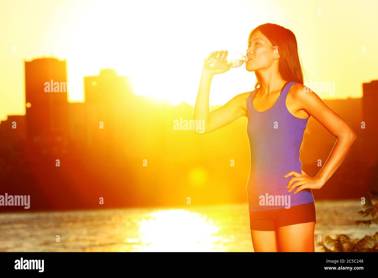 Femme buvant de l'eau après avoir fait du jogging. Une athlète féminine profite du soleil et se fait boire à la bouteille d'eau après avoir fait de l'exercice en plein soleil. Modèle de forme physique multiracial asiatique / caucasien fille Banque D'Images