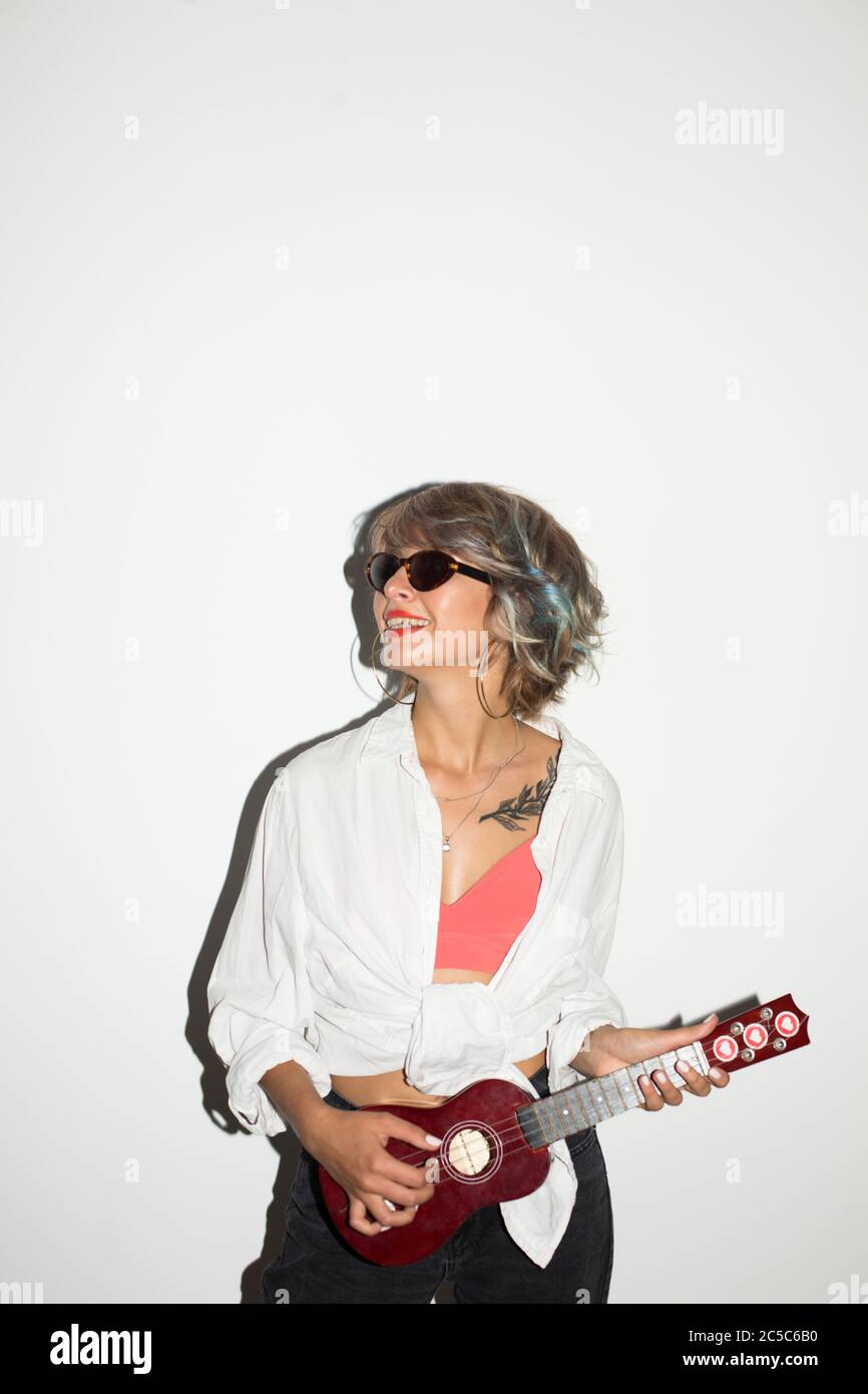 Cool sourire fille dans la chemise blanche et lunettes de soleil regardant de côté en jouant sur la petite guitare sur fond blanc Banque D'Images