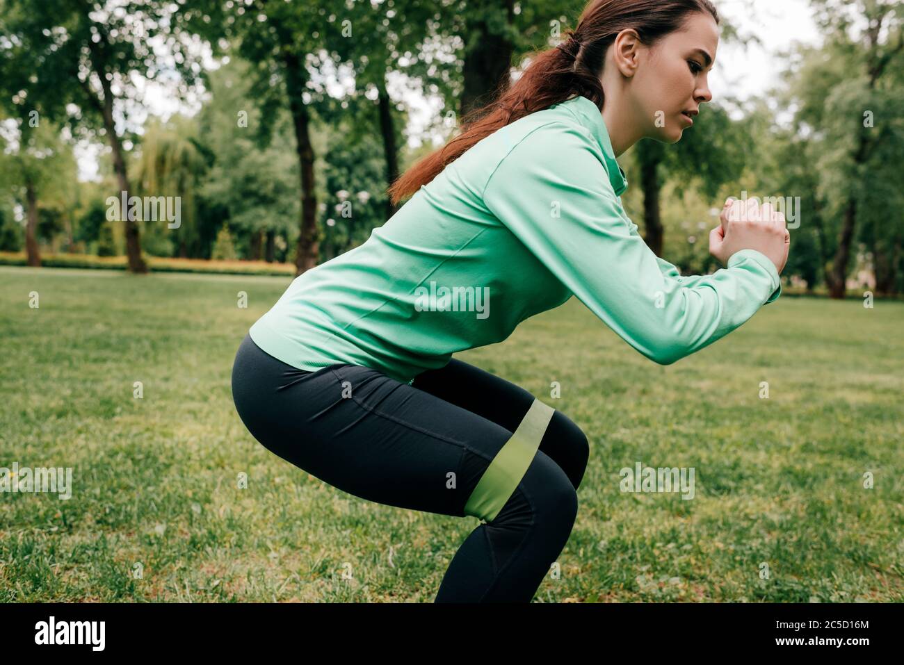 Vue latérale du sportswoman faisant du squat avec bande de résistance sur l'herbe dans le parc Banque D'Images