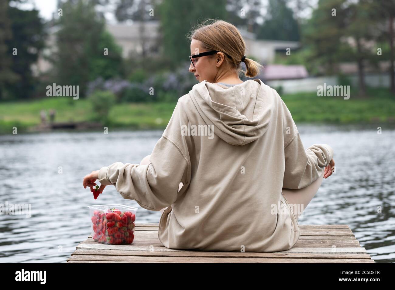 Jeune femme mangeant des fraises fraîches aux fruits de baies dans un récipient en plastique, reposant sur une jetée en bois de rivière, vue arrière. Heure d'été. La joie du pays lif Banque D'Images